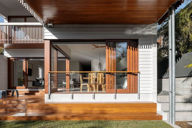 Exterior rear elevation of a contemporary multi-level home, focused on a covered timber-lined outdoor deck and balcony. The facade combines white horizontal weatherboard cladding, warm timber decking and soffits, large glazed openings, timber-framed sliding doors, and a metal cable balustrade. Interior dining and living areas are visible through the open glazing, with a landscaped lawn and side walkway in the foreground.