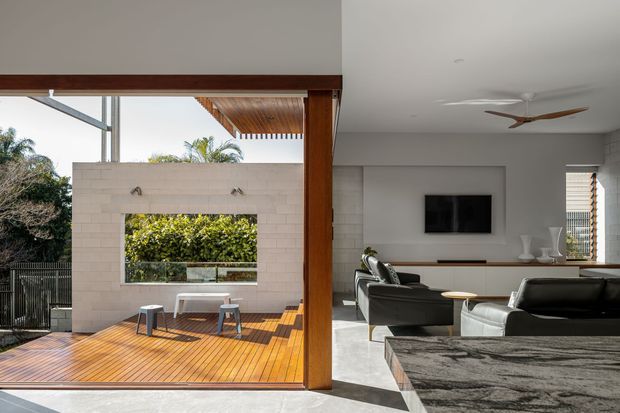 Open-plan contemporary living room opening onto a timber outdoor deck through large glazed sliding doors. The interior has white walls, a dark low-profile sofa, a wall-mounted television, built-in white cabinetry, and a sculptural ceiling fan. Outside, the deck includes a pale rendered brick privacy wall with a rectangular opening, outdoor seating, and integrated greenery. Strong indoor-outdoor connection with warm timber detailing and abundant daylight.