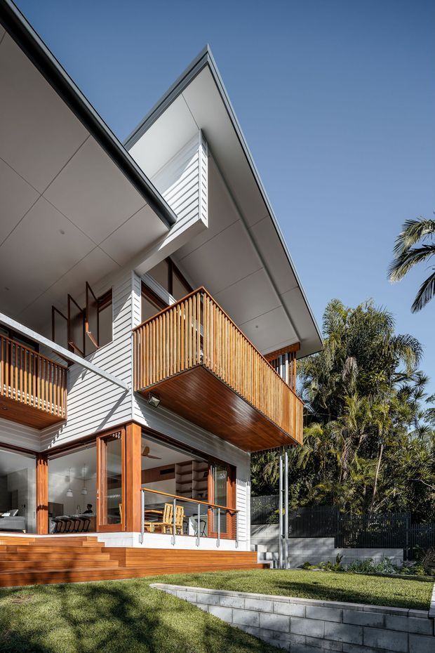 Exterior architectural photograph of a contemporary multi-level home with a sharply pitched roof, white horizontal weatherboard cladding, broad white soffits, timber-framed glazing, and projecting timber balconies. The balconies feature warm vertical timber balustrades and timber soffit lining, while stairs and a lawn lead up to the house. Tropical planting and a clear blue sky surround the property.