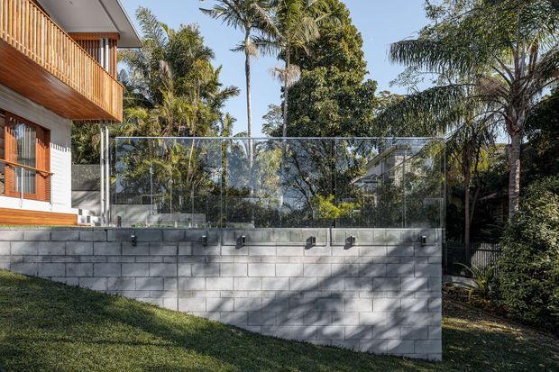 Exterior view of a contemporary multi-level residential property with a timber-clad balcony and window surrounds at left. A rendered light wall sits above a rendered block retaining wall, topped by a clear glass balustrade or pool barrier. Dense tropical landscaping and palms surround the structure, with strong sunlight and shadows across the wall and lawn.