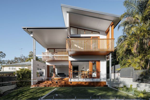 Exterior view of a contemporary multi-level home with dramatic asymmetrical skillion roof forms, broad eaves, white cladding, timber posts and decking, and prominent vertical timber balustrades. Covered outdoor living areas and glazed openings connect the house to a landscaped front yard, with mature palms and neighbouring residential buildings visible around it.