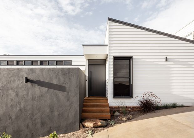 Exterior view of a contemporary single-storey dwelling with white horizontal weatherboard cladding, a sharply sloping roofline, dark-framed windows and door, and a textured rendered boundary wall. Timber entry steps lead to a recessed front door, with brickwork visible at the base and minimalist landscaping along the facade.