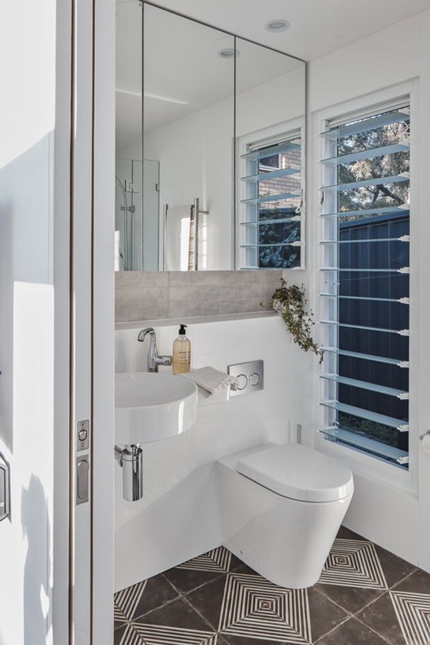 Bright compact contemporary bathroom viewed through an open doorway. The room features a wall-mounted white basin with chrome tap and exposed chrome waste, a wall-hung white toilet, a large mirrored cabinet or mirror wall, pale grey rectangular wall tiles, white walls, plantation-style louvre windows, and a bold dark geometric patterned floor tile. Natural daylight creates a clean, high-contrast atmosphere.
