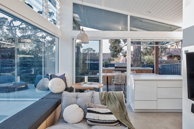 Bright contemporary living room with floor-to-ceiling glazing, a low sectional sofa, pendant light, and white built-in cabinetry. The glazing opens visually onto a covered outdoor entertaining area with a bar counter and stools. White-painted timber ceiling boards and large white-framed windows create an airy indoor-outdoor connection.