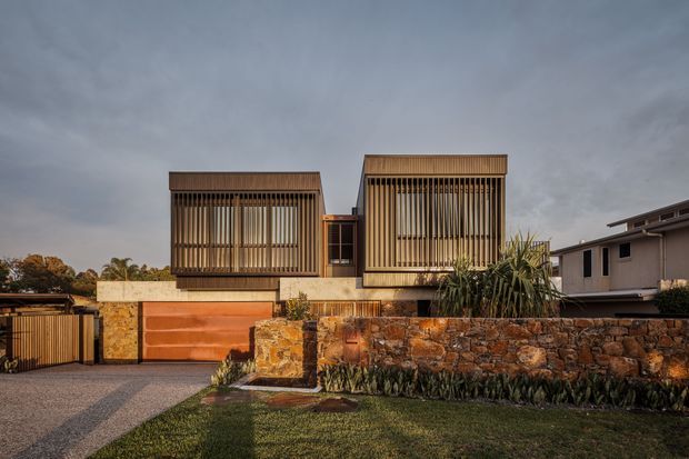 Contemporary two-storey residential facade with two prominent upper-level volumes screened by vertical timber battens. The house combines warm timber screening, rendered concrete elements, orange-toned garage doors, and rough natural stone retaining walls, with a landscaped front yard and driveway. The composition has strong horizontal and geometric lines with a flat or very low-pitched roof form.