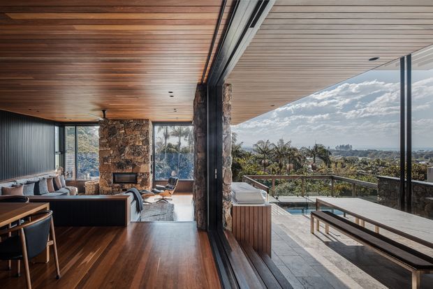 A refined indoor-outdoor living space with a timber-lined ceiling and warm timber flooring, opening through expansive glazing to a paved terrace with outdoor dining furniture and a swimming pool. Stone-clad walls, dark structural framing, and a panoramic leafy coastal or suburban outlook create a strong connection between the living room and terrace.