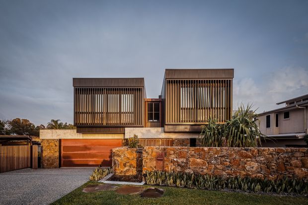 Front exterior view of a contemporary multi-level residence with two prominent box-like upper volumes screened by closely spaced vertical timber battens. The home combines warm timber detailing, exposed natural stone walls, concrete elements, and a large orange-toned garage door, with landscaped planting and a low stone boundary wall in the foreground.