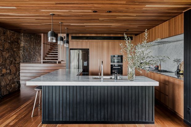 A sophisticated contemporary kitchen dominated by a large stone island with a black vertical-slat base, warm timber cabinetry and ceiling, integrated stainless-steel appliances, and dark pendant lighting. A rugged natural stone wall and open staircase add texture at the rear, while an expansive veined stone splashback and benchtop create a refined focal point.