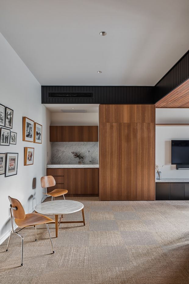 A contemporary open-plan living and dining area with a compact kitchen concealed behind warm timber cabinetry. The room features a round dining table with wooden chairs, patterned carpet flooring, white walls, a black vertical-panel feature bulkhead, a marble-look splashback, and a television wall with dark built-in cabinetry. A timber-lined ceiling section adds warmth and continuity to the joinery.