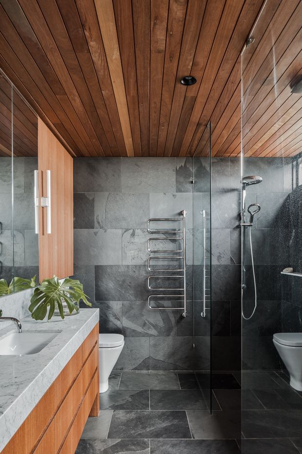 A contemporary bathroom with a walk-in glass shower, dark grey stone-look wall and floor tiles, a wall-mounted chrome towel warmer, and a timber-lined ceiling. The foreground features a marble vanity benchtop with an integrated basin and warm timber cabinetry. A toilet and shower fixtures are visible, with a large leafy plant adding natural contrast.