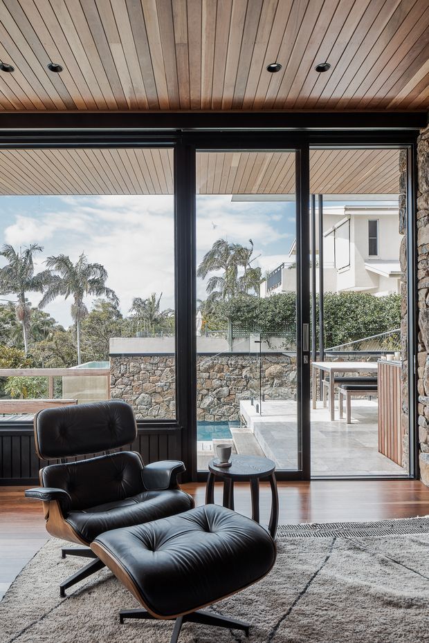 A refined living room opens directly onto a paved outdoor terrace and pool area through large black-framed glazed doors. The room features a timber-lined ceiling with recessed downlights, dark timber flooring, a rough natural-stone feature wall, and a black leather lounge chair with matching ottoman positioned over a large neutral rug. Outside, stone retaining walls, glass balustrades, palms, and neighbouring residential buildings are visible.