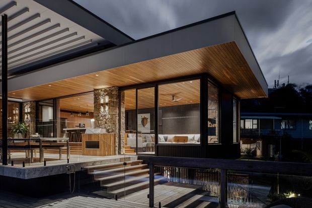 Dusk exterior view of a contemporary multi-level residence with extensive floor-to-ceiling glazing, dark metal framing, a broad flat roof and warm timber soffits. Stone-clad columns, timber decking, outdoor stairs and a reflective pool or water feature frame the open living and kitchen spaces, which are visible through the glass. A covered pergola structure extends at the upper left.