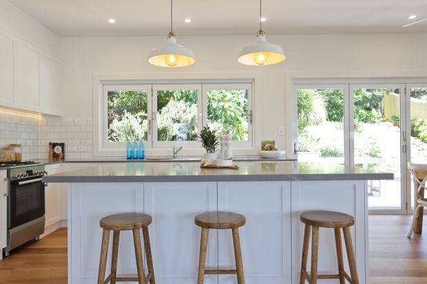 Bright, spacious white kitchen with a large island and three timber bar stools. The room features white Shaker-style cabinetry, a pale stone benchtop, timber flooring, tiled splashback, pendant lighting, and generous windows and glazed doors opening to greenery outside.