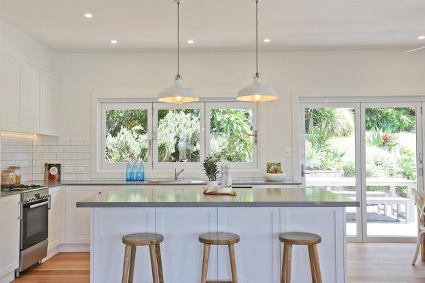 Bright, spacious white kitchen with a central island and three timber bar stools. The room features white shaker-style cabinetry, white subway-tile splashback, pale timber flooring, a grey benchtop, stainless-steel appliances, pendant lights, and generous windows and glazed doors opening to a leafy outdoor area.