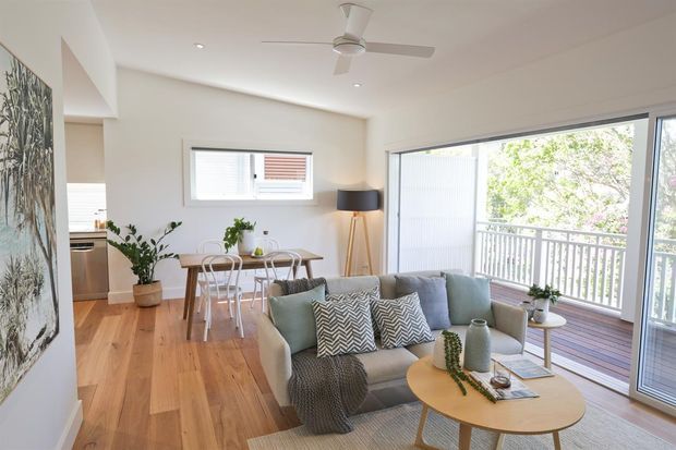 Bright open-plan living and dining area with warm timber flooring, white walls, a pale upholstered sofa, round timber coffee table, compact dining setting, indoor plants, and extensive glazed doors opening to a balcony. A small kitchen is partially visible at the rear.