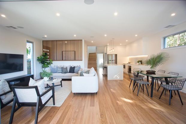 Bright open-plan living, dining, and kitchen interior with light timber flooring, white walls, a white sectional sofa, black-framed dining furniture, and a compact white kitchen island. A timber vertical-slat feature screen separates the living area from the hallway, while large windows and recessed lighting create an airy contemporary atmosphere.