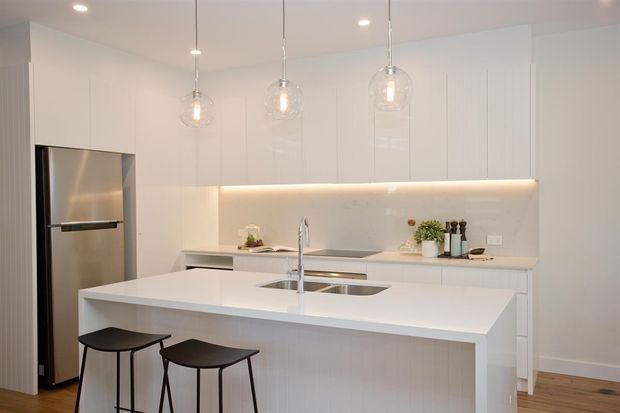 Bright contemporary white kitchen with a large island and integrated sink, two black stools, full-height white cabinetry, a stainless-steel refrigerator, pale benchtops, and a softly illuminated backsplash. Three clear glass pendant lights hang above the island, while timber flooring adds warmth.