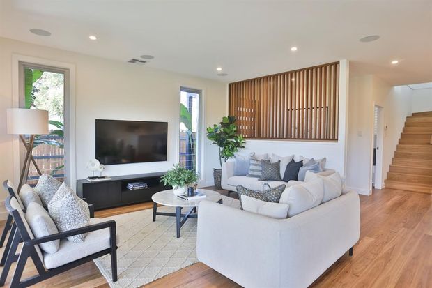 Bright contemporary living room with light timber flooring, a large pale sectional sofa, patterned armchair, round coffee table, wall-mounted television, indoor plant, and a striking vertical timber-slat feature screen. An open stairway and hallway are visible to the right, with recessed ceiling lighting and large windows providing natural light.