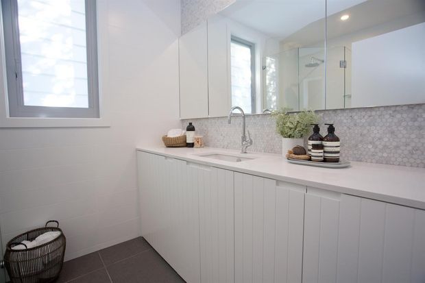 Bright, contemporary bathroom with a long white vanity featuring vertical-panel cabinetry, an integrated basin and chrome mixer tap. A pale hexagonal mosaic tiled splashback runs beneath mirrored wall cabinets, with a glass-enclosed shower reflected in the mirror. White wall tiles, a small window, recessed ceiling lighting, and dark floor tiles complete the clean, light-filled space.