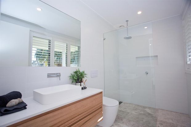 Bright, contemporary bathroom with a floating timber vanity, white vessel basin, wall-mounted tapware, concealed-cistern toilet, and frameless glass walk-in shower. White tiled walls, pale stone-look floor tiles, plantation shutters, and recessed lighting create a clean, airy interior.