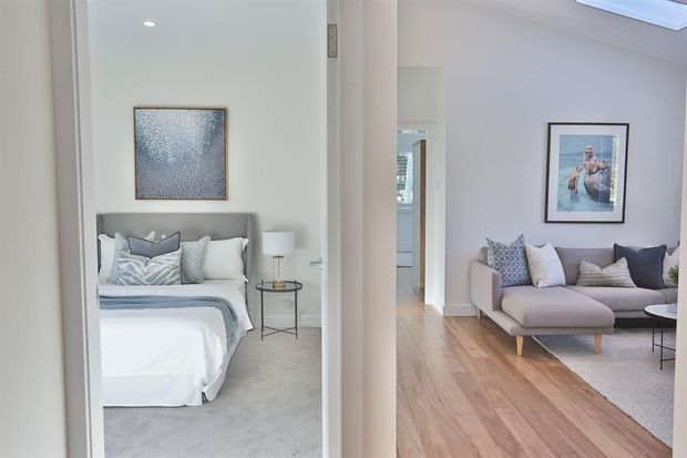 A bright, refined view through an interior opening showing a bedroom on the left and a living room on the right. The bedroom has a neatly made upholstered bed, soft grey bedding, a small round side table and abstract wall art, while the living room contains a pale upholstered sectional sofa, patterned cushions, framed artwork and light timber flooring. The palette is predominantly soft white, grey, beige and light wood.