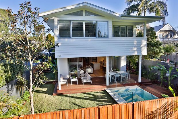 A bright two-storey coastal-style home viewed from the garden, featuring a white horizontal weatherboard upper level with a front-facing gable roof, extensive louvred and glazed openings, and an open lower-level living and dining area. A timber deck, small swimming pool, landscaped garden, and timber privacy fence occupy the foreground.