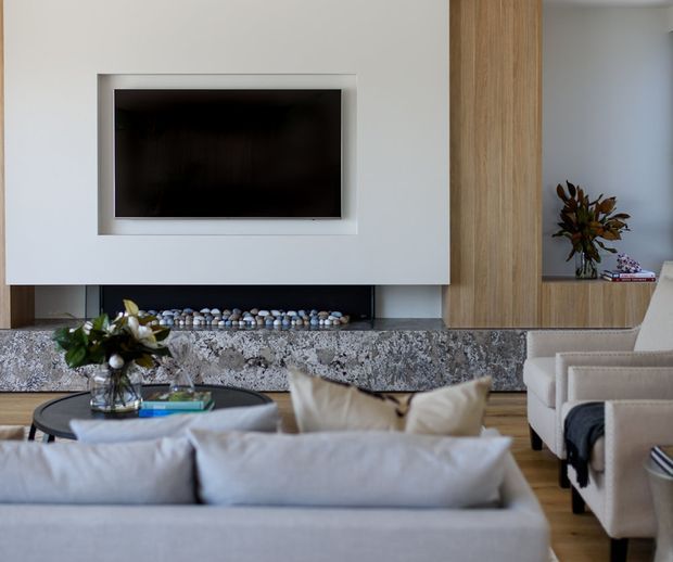 Contemporary living room centered on a wall-mounted television above a long stone-clad fireplace hearth with a pebble-filled firebox. Floor-to-ceiling timber-toned built-in cabinetry frames the feature wall, while a pale upholstered sofa, armchairs, round coffee table, and restrained decorative styling create a refined neutral interior.