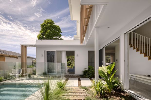 A bright contemporary courtyard-style residential exterior with a swimming pool in the foreground, landscaped planting, stepping-stone paving, and extensive white-framed glazing. The view looks toward a white multi-level home with a covered outdoor living area and a visible internal staircase behind open glazing. Timber-toned vertical screening and soffit detailing add warmth to the crisp white facade.