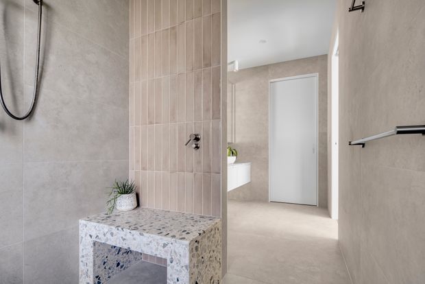 A contemporary bathroom featuring a walk-in shower with large light-grey wall tiles, a vertical blush-beige tiled feature wall, and a built-in terrazzo bench with an open base. A frameless glass shower screen separates the shower area from the rest of the room. The adjoining vanity area and a closed white door are visible in the background. Black shower hardware and a black towel rail provide contrast against the neutral finishes.