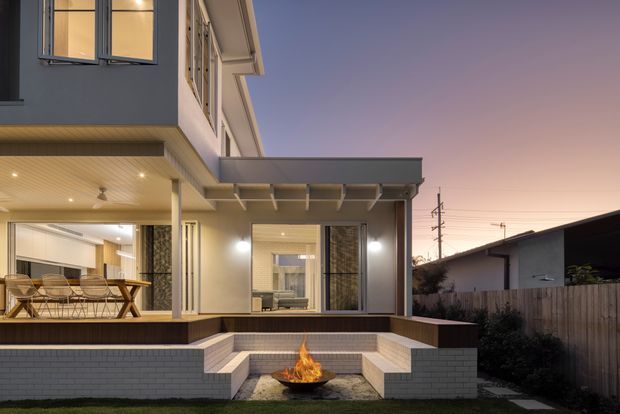 Twilight exterior view of a contemporary multi-storey home with a covered patio and outdoor lounge area. The house features dark and light rendered wall surfaces, extensive glazing, timber-look soffit lining, a projecting flat roof, and a stepped paved terrace with a built-in outdoor fire bowl. A neighbouring structure and timber boundary fence are visible at the side.