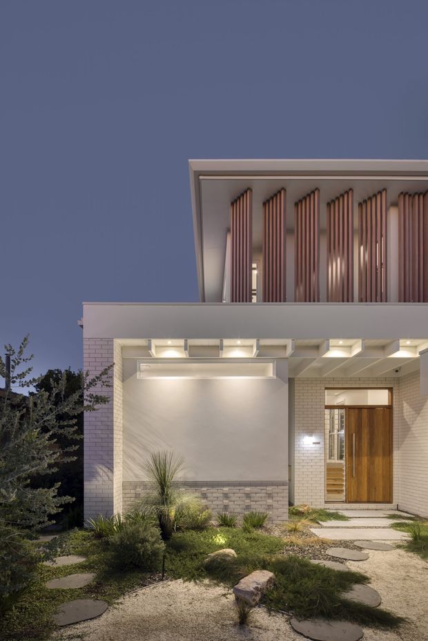 Dusk-time exterior photograph of a contemporary two-storey home facade. The light-coloured brick and rendered entry elevation features a broad projecting canopy, recessed downlighting, a timber front door, and an upper-level screened balcony or facade with repeated vertical timber battens. A drought-tolerant landscaped front garden with gravel and stepping stones leads to the entry.