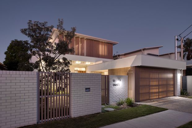 Twilight street-level view of a contemporary multi-level residence with a white rendered and brick facade, strong horizontal rooflines, vertical timber-toned screening, and an integrated double garage. A masonry front wall and slatted pedestrian gate enclose a landscaped entry garden, while warm interior lighting creates a welcoming glow.