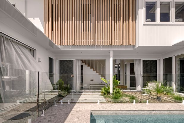 A bright contemporary multi-level residential courtyard viewed from beside a swimming pool. White rendered walls, extensive glazing with dark frames, glass balustrades, pale stone paving, and a prominent upper-level screen of vertical timber battens define the architecture. An internal stair and landscaped courtyard are visible beneath the overhanging upper floor.