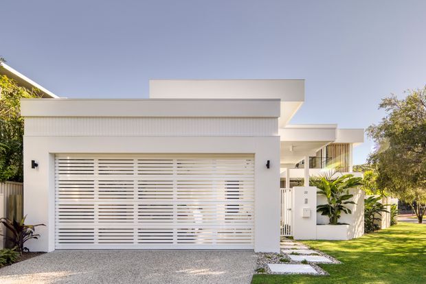 Bright contemporary residential facade viewed from the street, featuring a white rendered garage volume with a horizontal slatted garage door, layered flat roof forms, white entry fencing, a stepping-stone path, and landscaped garden surrounds.