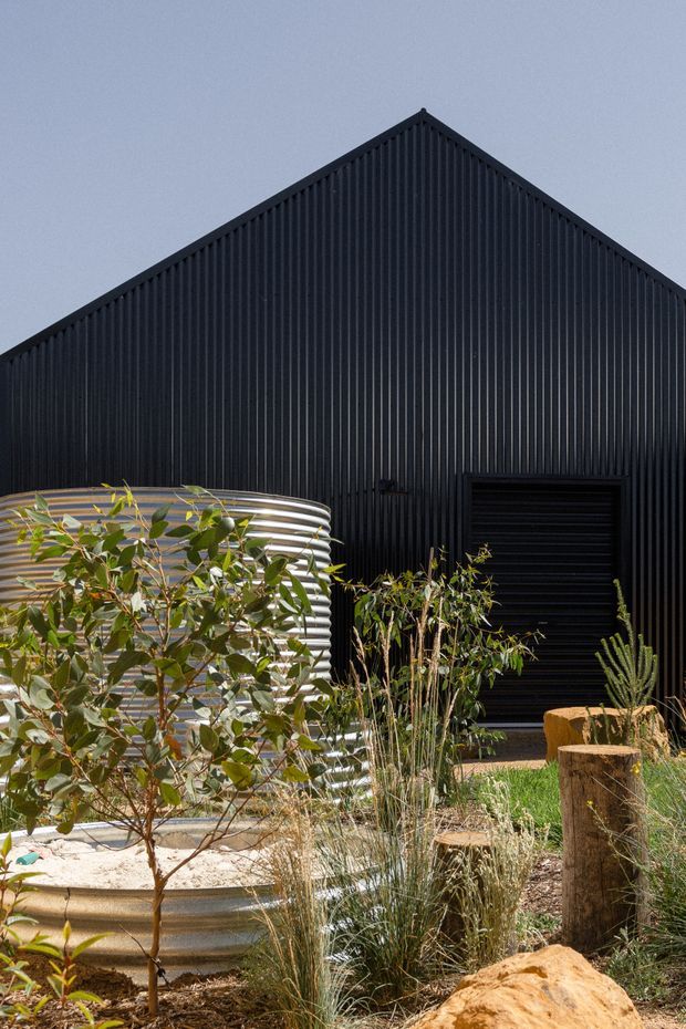 Exterior view of a dark corrugated metal gable-roof building with a black roller door and a prominent silver corrugated cylindrical water tank in a landscaped native-style garden. Drought-tolerant grasses, shrubs, timber posts, and a large natural rock occupy the foreground.