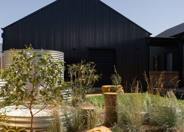 Exterior view of a dark, corrugated-metal-clad gabled building with a smaller adjoining wing and recessed entry. The foreground features a landscaped garden with ornamental grasses, shrubs, timber stump elements, and a large corrugated metal cylindrical water tank or garden feature.