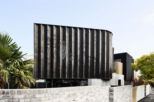 Contemporary multi-level residential building viewed from the street, featuring a striking curved facade of repetitive vertical fins with perforated dark screens, contrasting dark cladding, concrete elements, and a pale masonry boundary wall. Tropical planting frames the modern exterior.