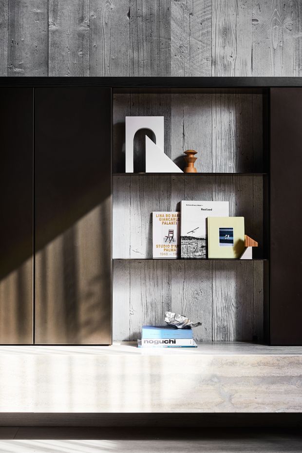 A close-up of a dark built-in storage and display unit with open shelving, set against vertically textured grey timber-look wall panels. The shelves hold books, sculptural objects and a small wooden vessel. Strong sunlight creates a diagonal shadow across the cabinetry and display, while a pale stone or concrete-look horizontal surface runs across the lower portion of the image. The composition is refined, monochromatic and material-focused.