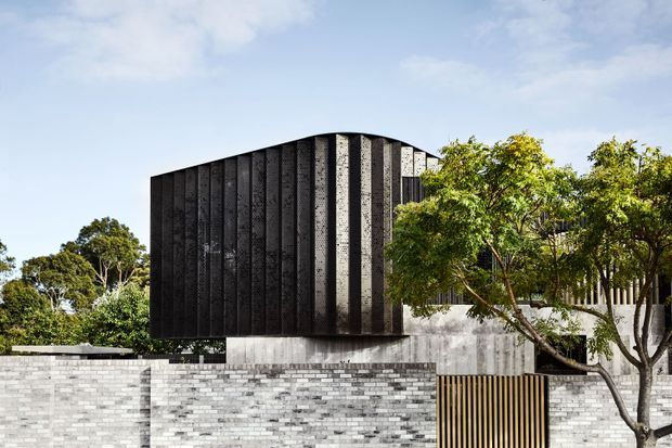 Exterior architectural photograph of a contemporary, multi-level building with a dark vertically ribbed and textured upper volume, pale concrete walls, and a light grey brick boundary wall. A timber-toned vertical slatted gate and mature trees frame the facade beneath a partly cloudy sky.