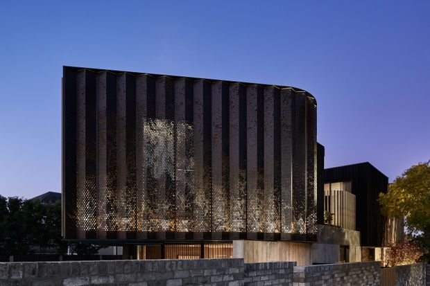 Contemporary multi-level building viewed externally at dusk, with a prominent dark perforated metal screen facade formed by deep vertical fins. Warm interior lighting glows through the screen and glazing, while a stone masonry boundary wall and landscaped trees frame the property.