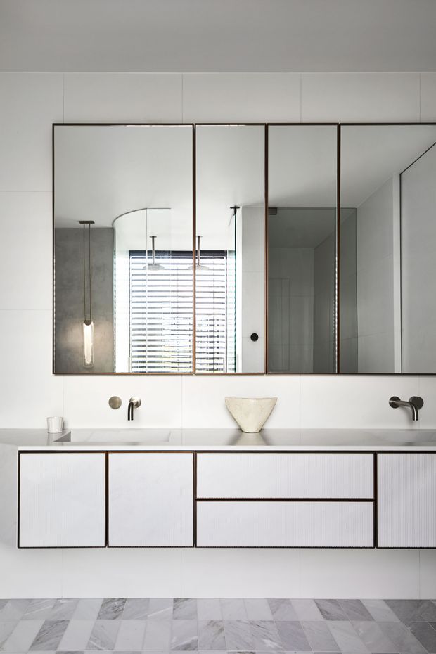 A minimalist contemporary bathroom vanity with a long floating cabinet, integrated countertop, two wall-mounted dark metal faucets, and a sculptural vessel basin. Large framed mirrored cabinet doors span the wall above the vanity, reflecting a window with horizontal blinds and a shower area. The room is finished in white wall panels and pale veined marble-look floor tiles, with restrained dark trim detailing.