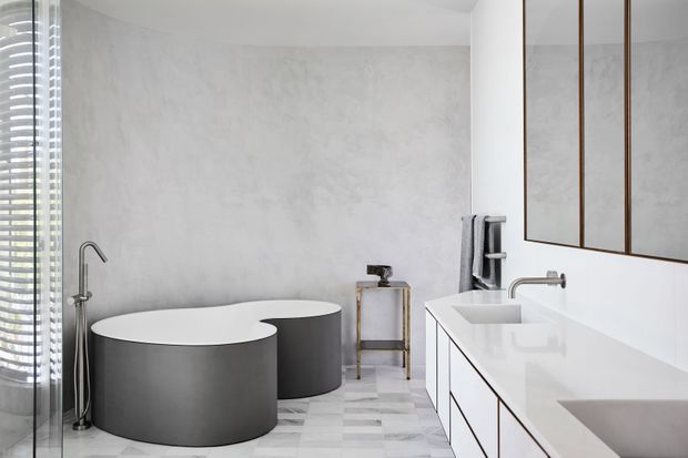 Contemporary minimalist bathroom featuring two sculptural freestanding matte-grey tubs, a large white double vanity with integrated basins, wall-mounted chrome tapware, full-height mirrored cabinetry with dark trim, and pale patterned stone or tile flooring. A textured light-grey plaster wall and sheer window provide a calm, monochromatic setting.