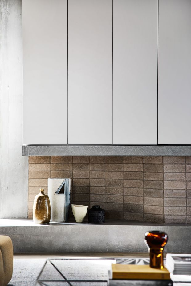 Close-up interior view of a contemporary kitchen or living space featuring tall white flat-panel cabinetry, a dark grey stone or concrete benchtop, and a brown-grey exposed brick backsplash. Minimal decorative ceramics sit on the counter, with part of a coffee table and seating visible in the foreground.