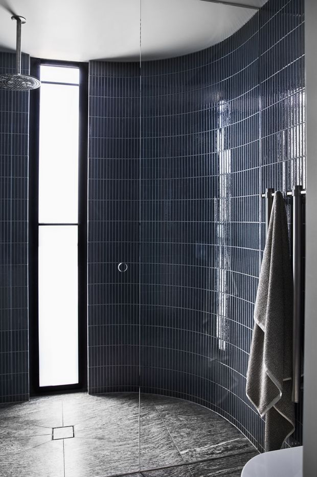 A dramatic contemporary bathroom with a curved wall clad in narrow glossy dark blue-black tiles, a frameless glass shower enclosure, a wall-mounted rainfall shower, and a tall frosted window. Large grey stone-look floor tiles and a dark towel rail reinforce the moody, refined palette.