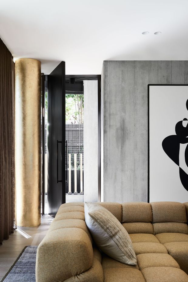 A refined contemporary living room with a large curved modular beige sofa in the foreground, a textured neutral cushion, dark timber wall panelling, a tall rounded metallic-gold feature column, and a grey vertically textured concrete-look wall. A black-framed glazed door opens toward an exterior balcony or walkway, while a bold black-and-white artwork adds contrast.