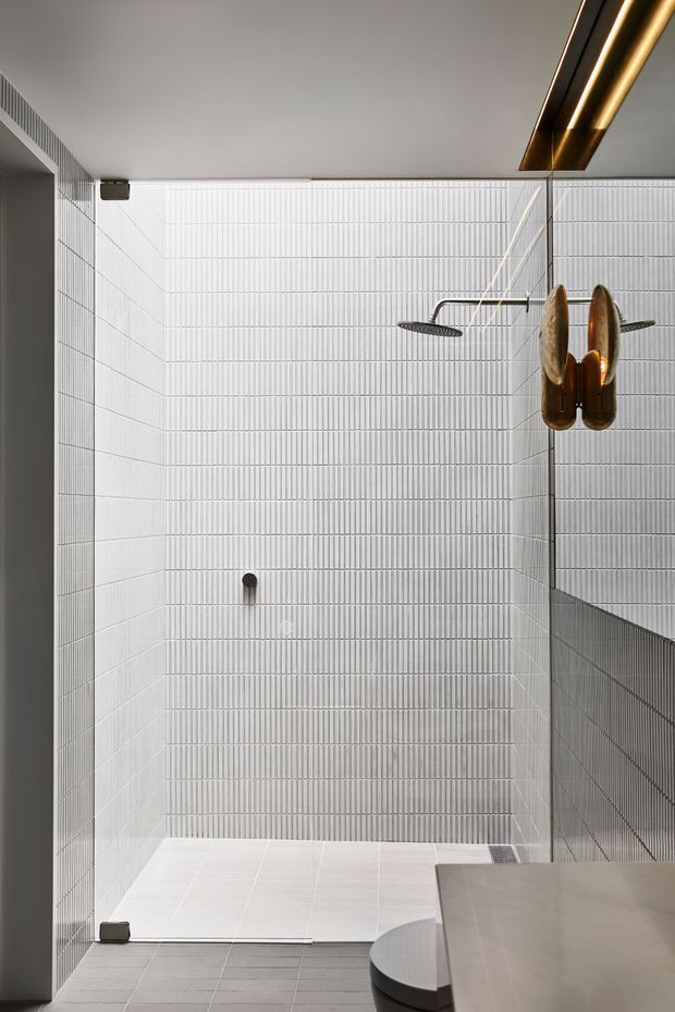 A minimalist walk-in shower with white vertically ribbed wall tiles, pale floor tiles, frameless glass screening, and sleek dark metal shower fittings. A sculptural bronze pendant light hangs beside a pale stone or concrete vanity edge, creating a refined contemporary bathroom interior.
