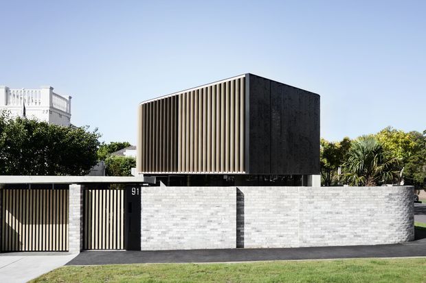 Street-facing exterior view of a contemporary two-storey residence. A dark rectangular upper volume with closely spaced vertical fins projects above a pale grey masonry boundary wall and matching gated entry, creating a strong screen-like facade. Tropical and suburban landscaping surrounds the property.