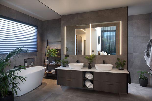 A contemporary, spacious bathroom with a freestanding white bathtub, double floating vanity, two vessel basins, large illuminated mirror, and dark grey stone-look wall and floor finishes. A window with horizontal blinds, open shelving, towels, and indoor plants complete the refined interior.