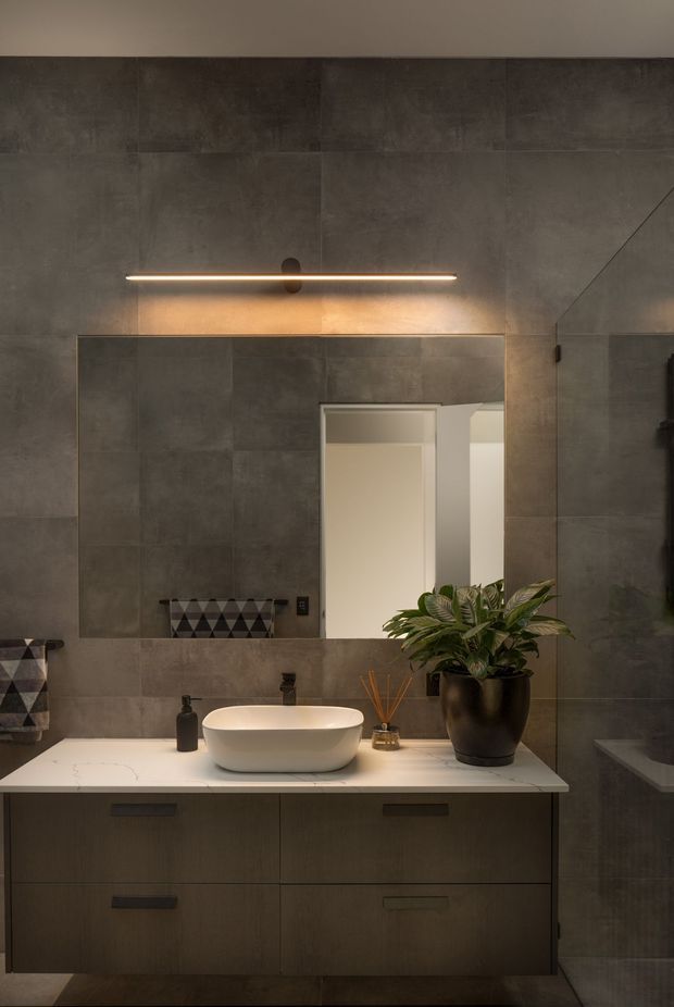 A moody contemporary bathroom vanity with large-format dark grey concrete-look wall tiles, a white vessel basin on a pale stone benchtop, dark timber cabinetry, black tapware, and a wide frameless mirror illuminated by a slim horizontal wall light. A potted plant and small accessories add warmth.
