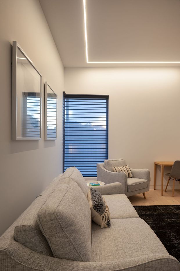 A softly lit contemporary living room with a large light fabric sofa in the foreground, a matching upholstered armchair, a small side table, and a compact desk and chair near the window. Neutral beige walls and upholstery are complemented by dark horizontal window blinds, framed artwork, a dark rug, and a recessed linear LED ceiling detail.